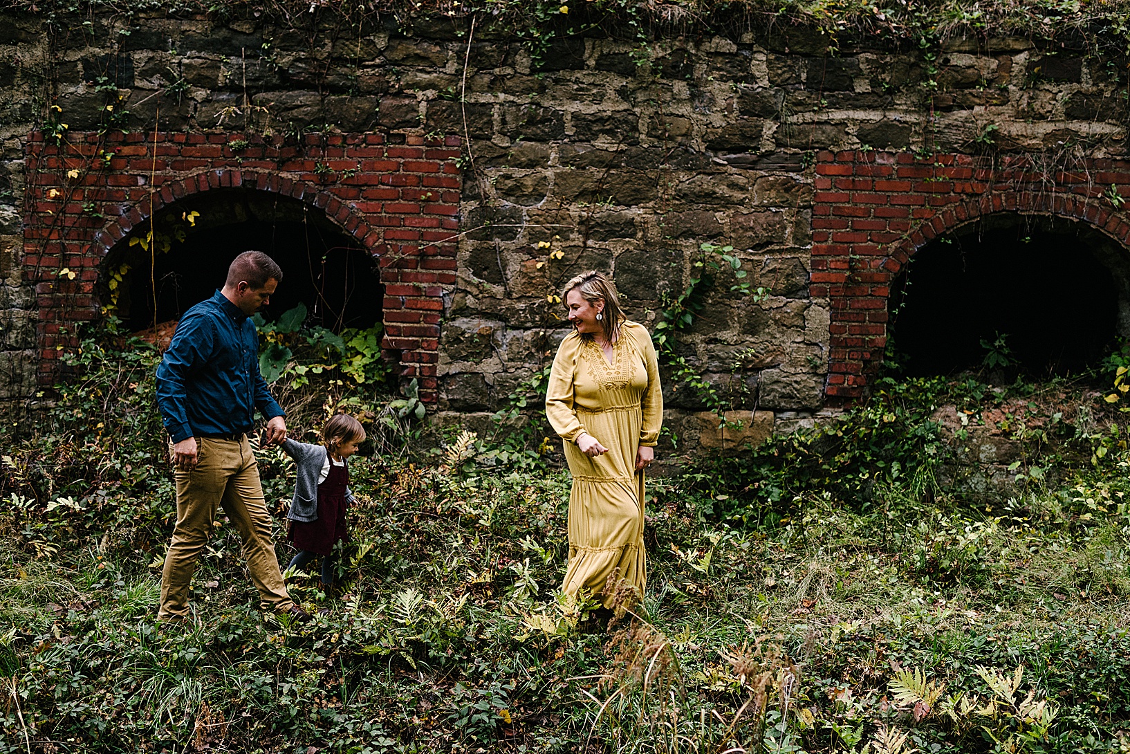 Blonde woman in mustard colored dress walks looking back at her daughter and husband holding hands walking behind her while in front of a gorgeous stone and brick wall at Leetonia Coke Ovens.