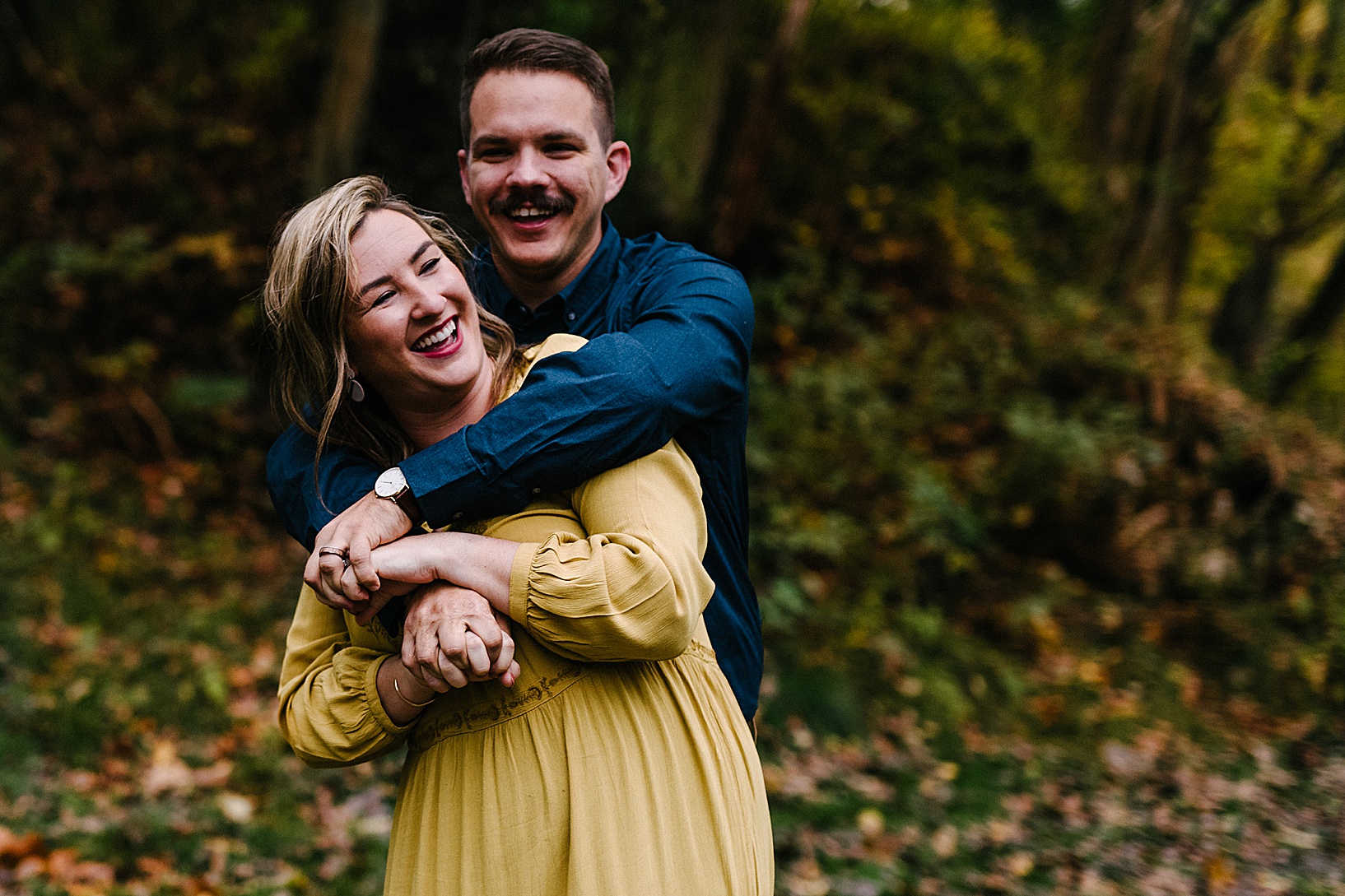 Blonde woman in mustard colored dress laughs and smiles while her mustached husband hugs her from behind with their fingers interlocked in front of them.