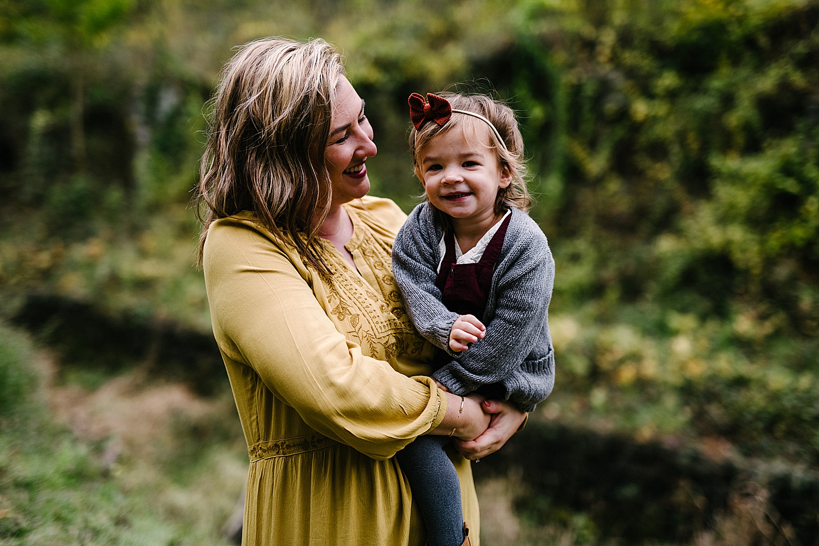 Blonde woman in a mustard dress smiles and looks at her daughter while she holds her in front of gorgeous greenery at the Leetonia Coke Ovens.