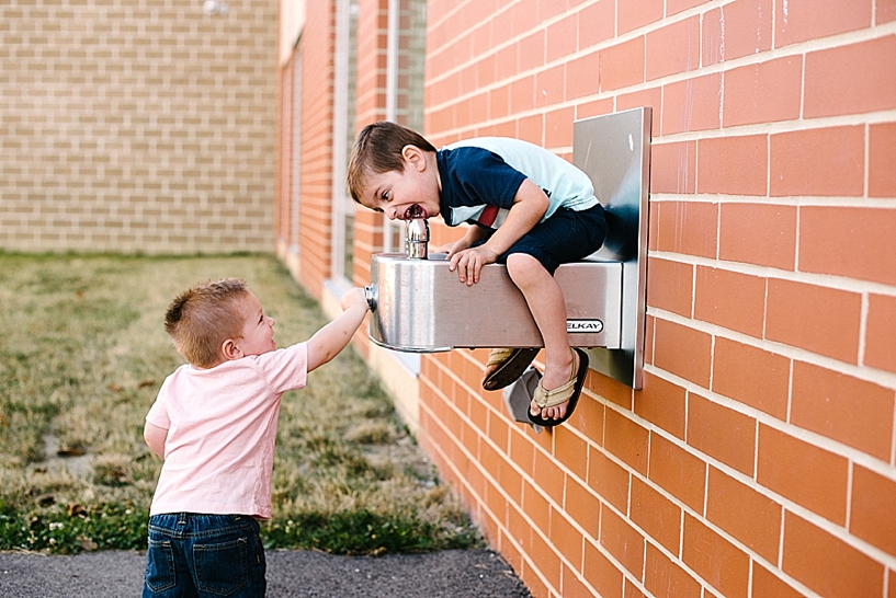 boy sitting on water fountain with brother helping push the button