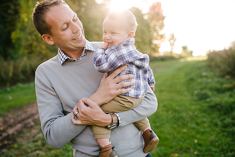 dad in grey sweater holding baby boy in plaid shirt in his arms with sun shining behind them