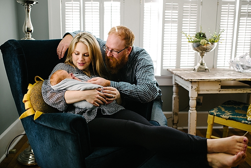 mother sitting on navy armchair holding newborn with husband's arms around them