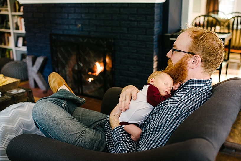 father with red hair and beard sitting by fireplace holding newborn son