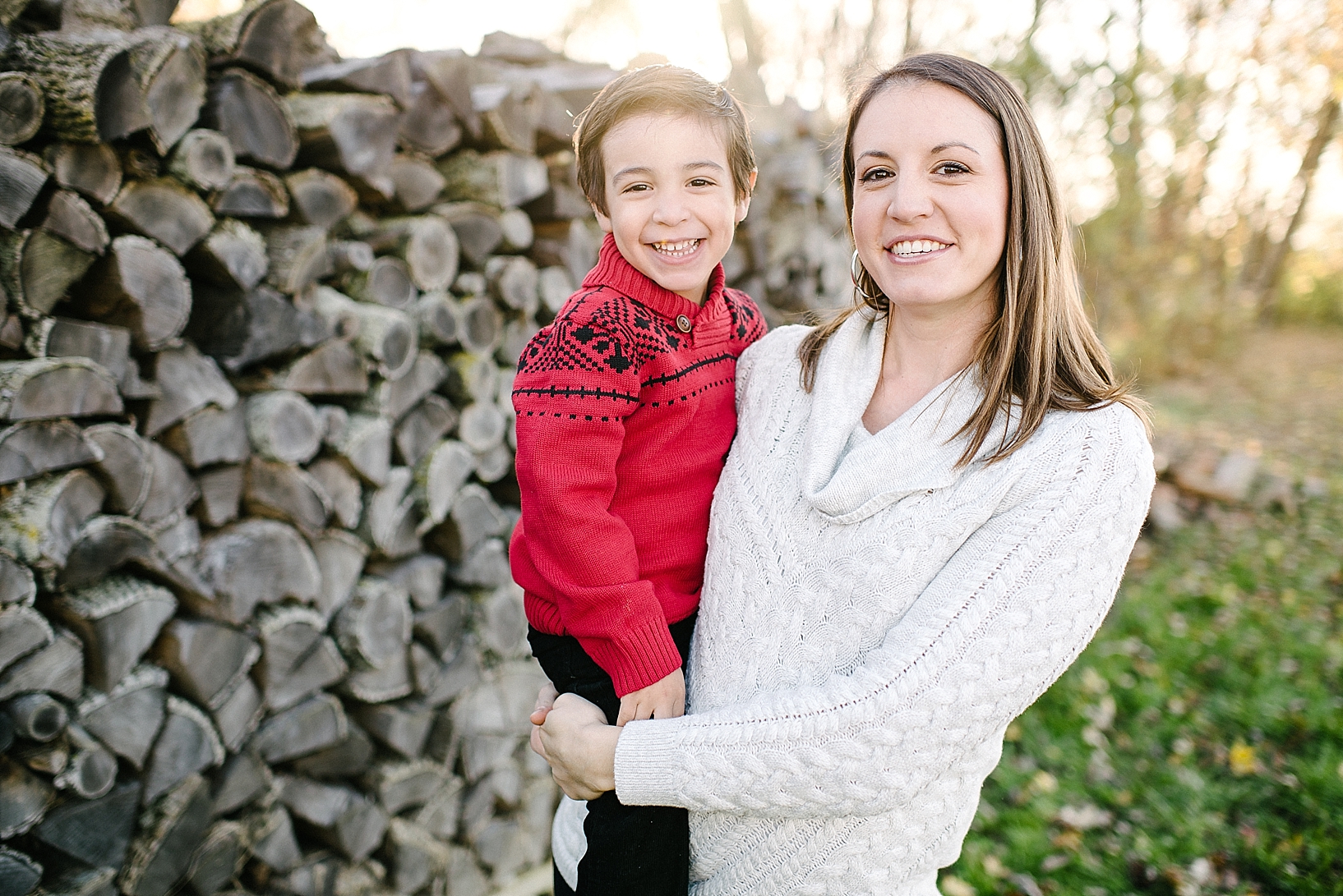 mom in cream sweater holding toddler son in red sweater standing in front of log pile