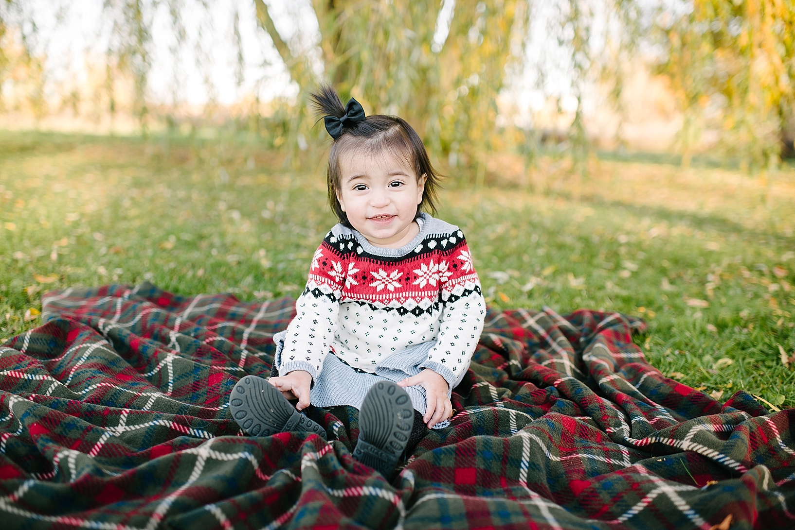 little girl in Christmas sweater and ponytail sitting on plaid blanket