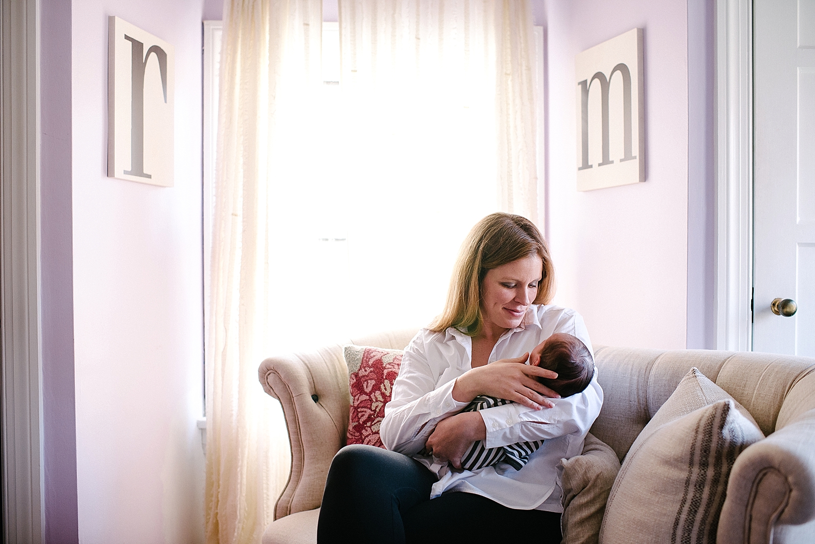 mother wearing white button down shirt sitting on chaise in front of window holding newborn baby