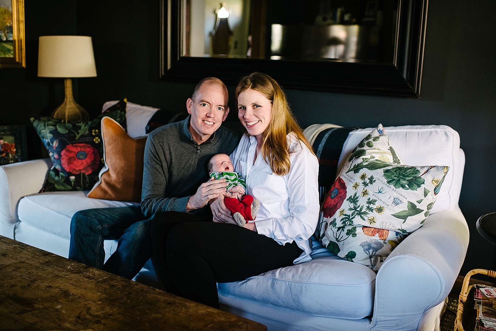 parents holding newborn sitting on living room couch smiling