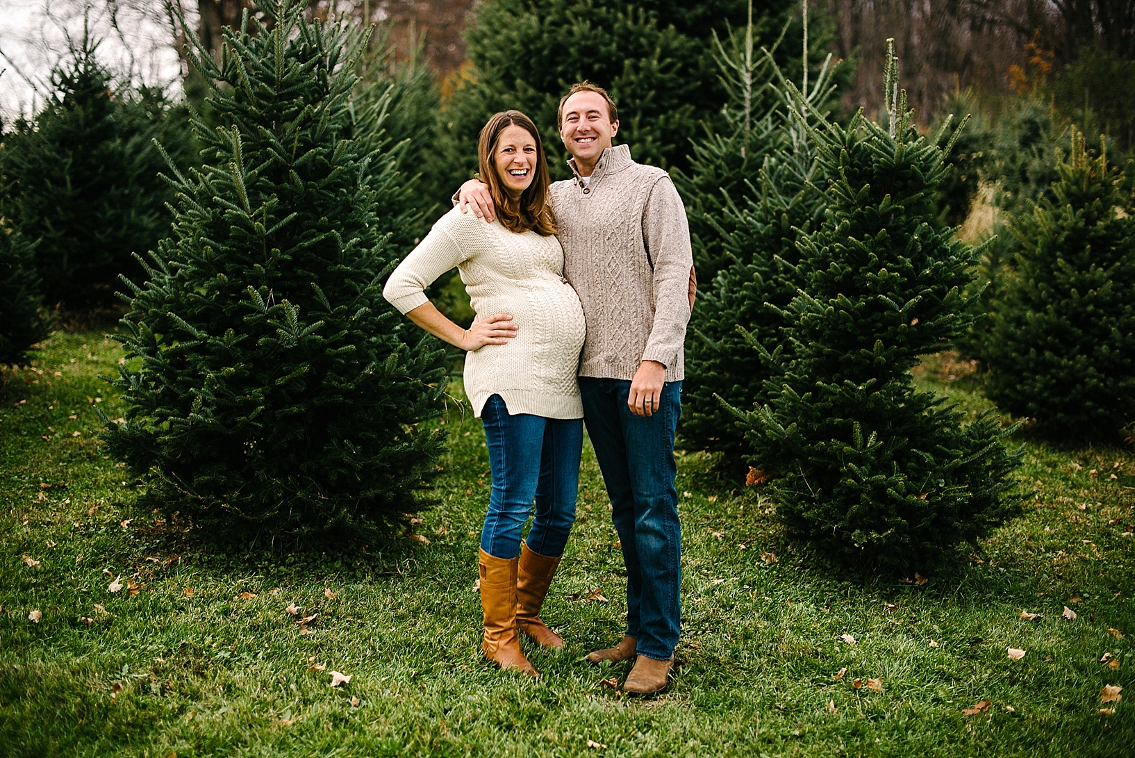 expectant mother and father wearing cable knit sweaters laughing among pine trees