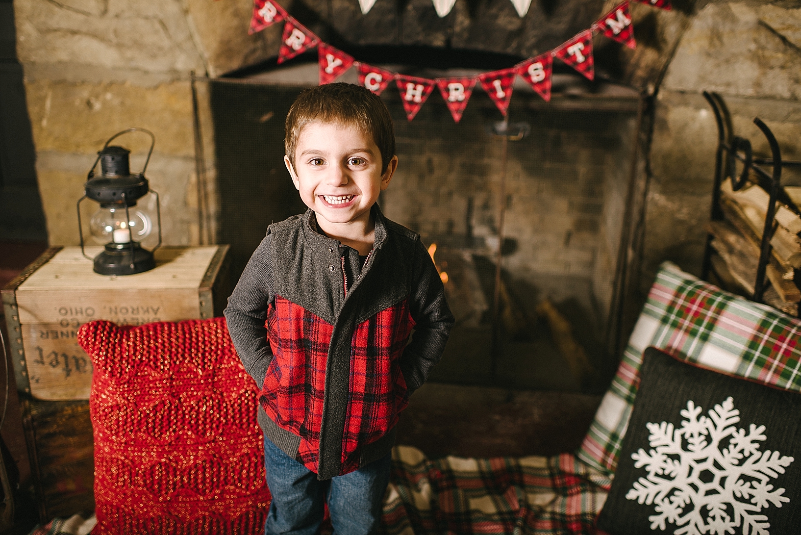 little boy wearing red and black flannel vest standing in front of fireplace smiling
