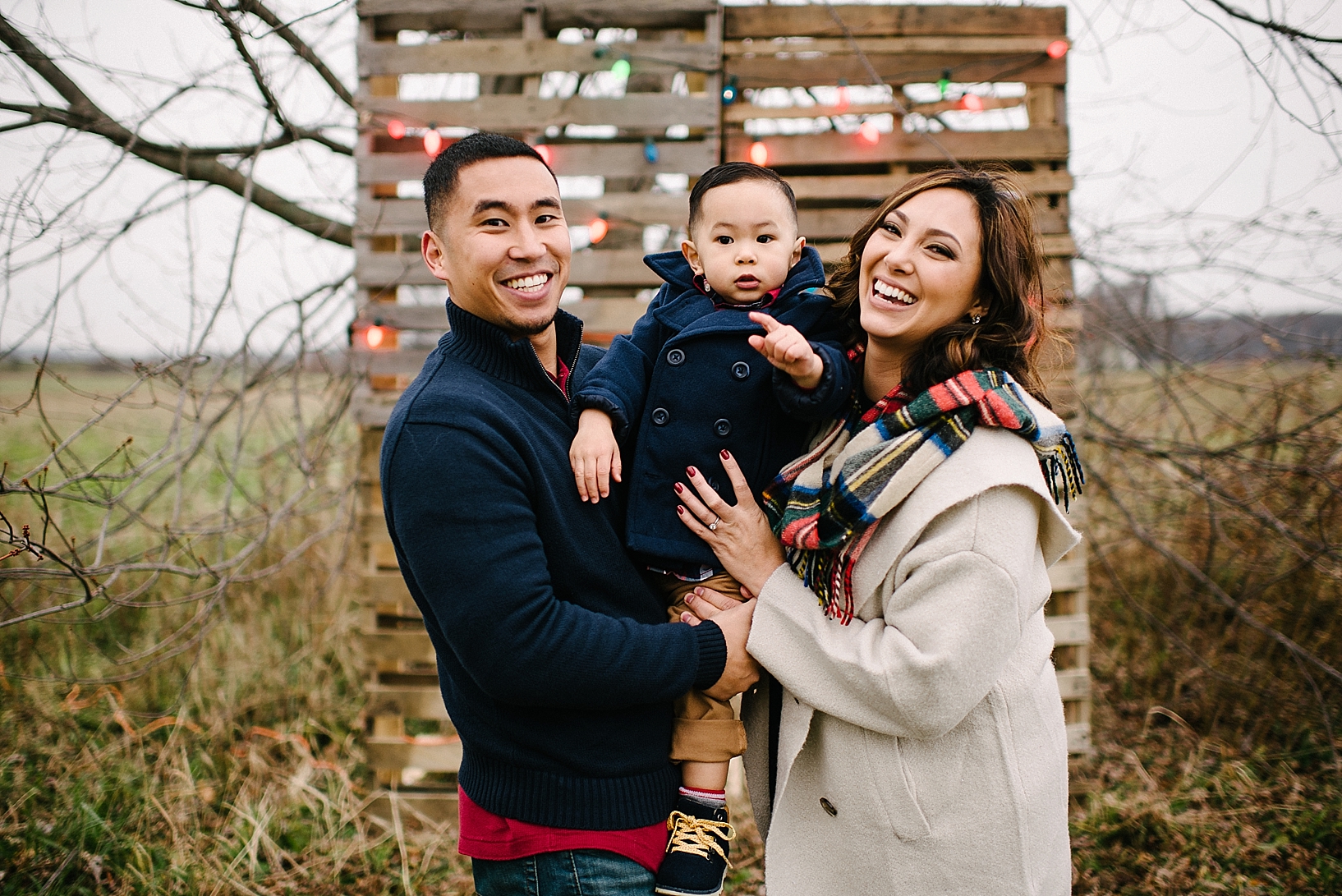 parents dressed in winter coats holding toddler son in navy pea coat outdoors in front of DIY wooden pallet wall with Christmas lights
