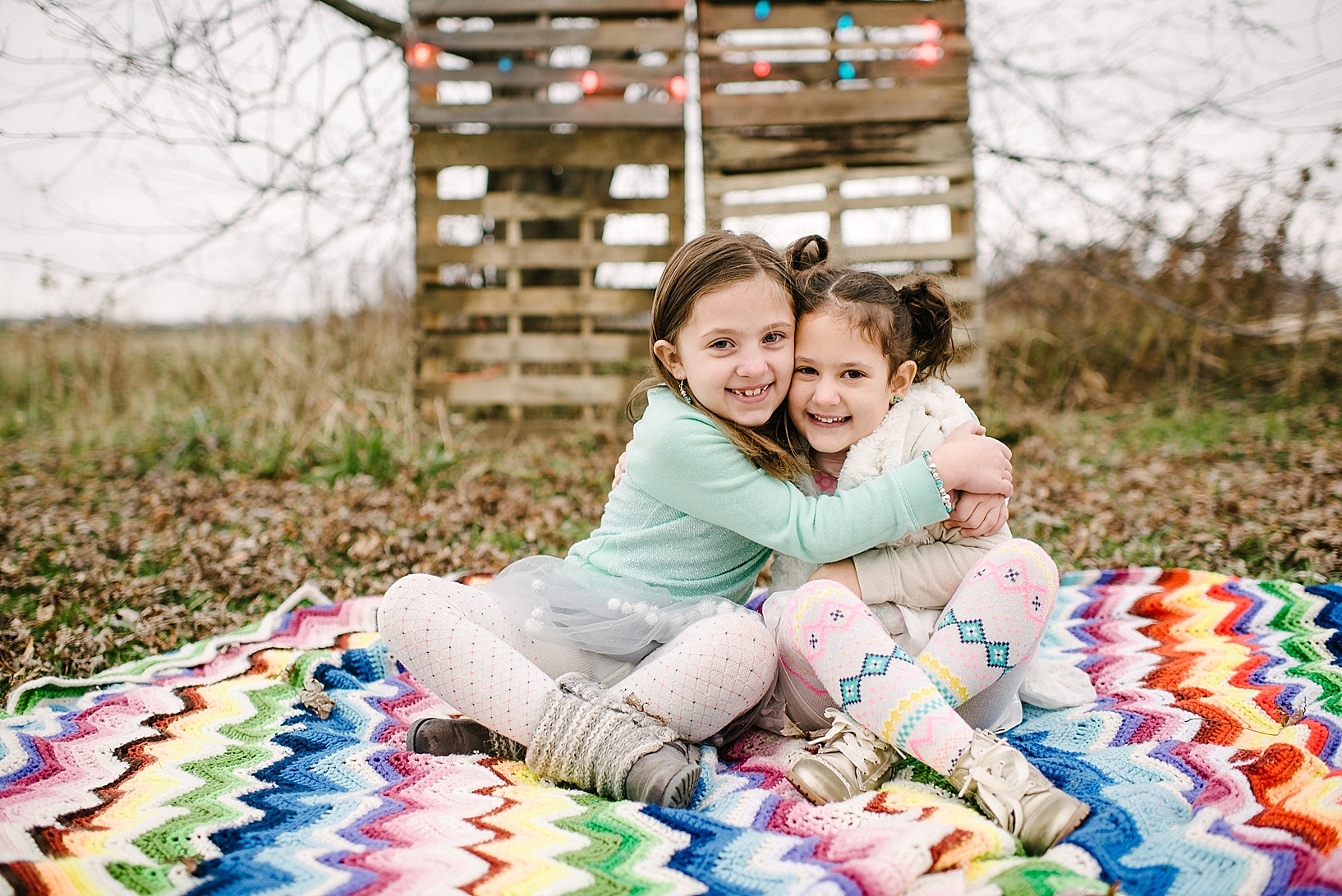 sisters sitting on colorful knit blanket hugging with Christmas lights in the background