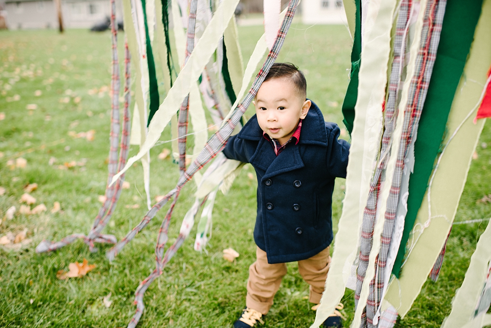 toddler boy wearing navy pea coat and khakis standing in curtain of streamers outside
