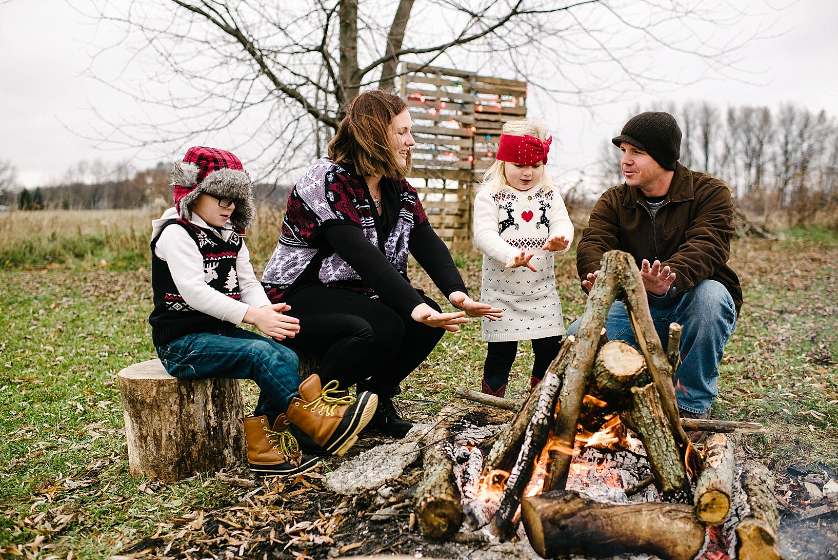 family sitting on tree stumps warming hands by campfire