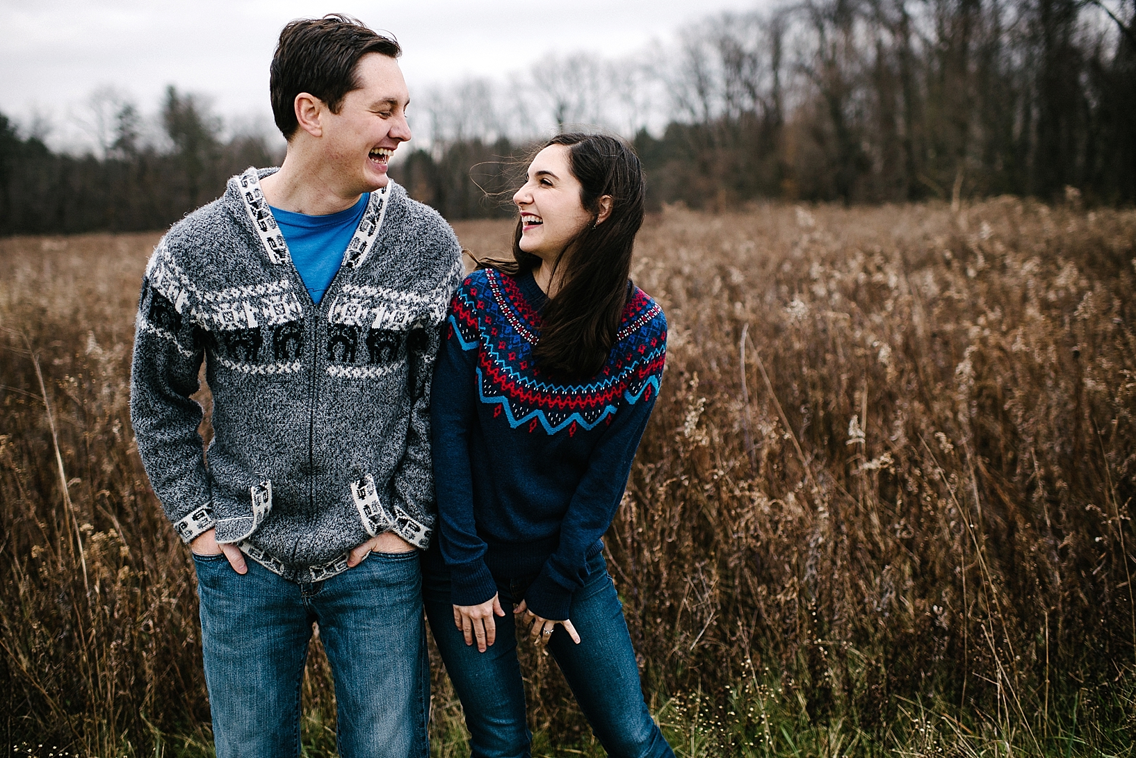 guy in grey sweater and girl in blue sweater standing in field laughing