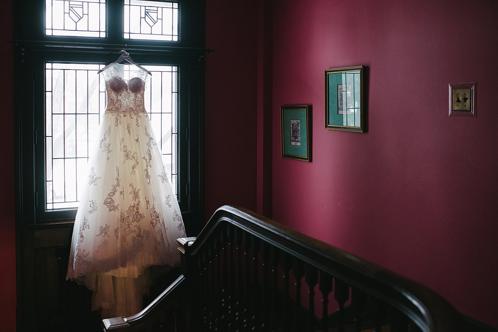 wedding dress hanging on window near old staircase and maroon walls
