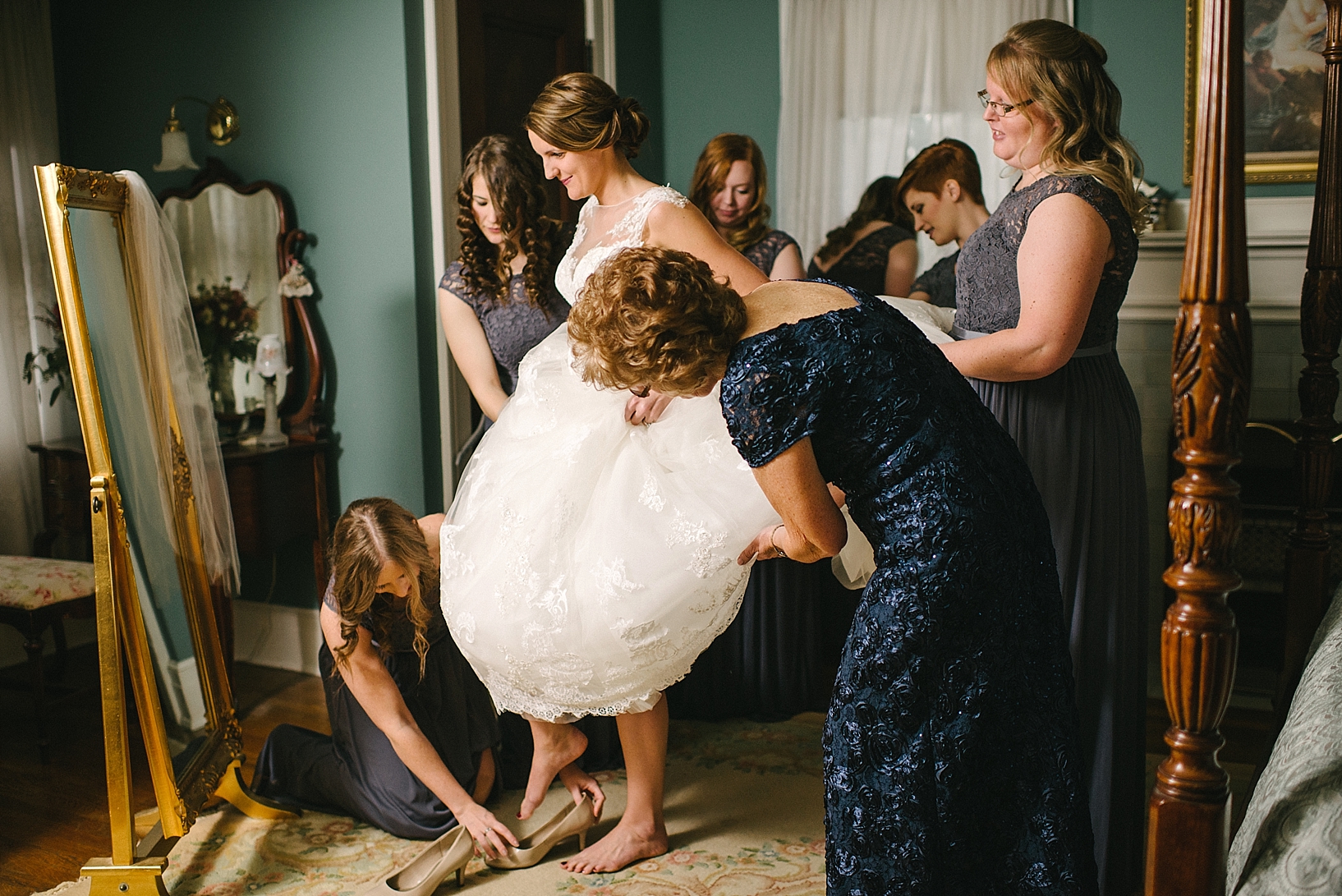 bridesmaid helps bride step into her shoes as bridesmaids hold her gown up