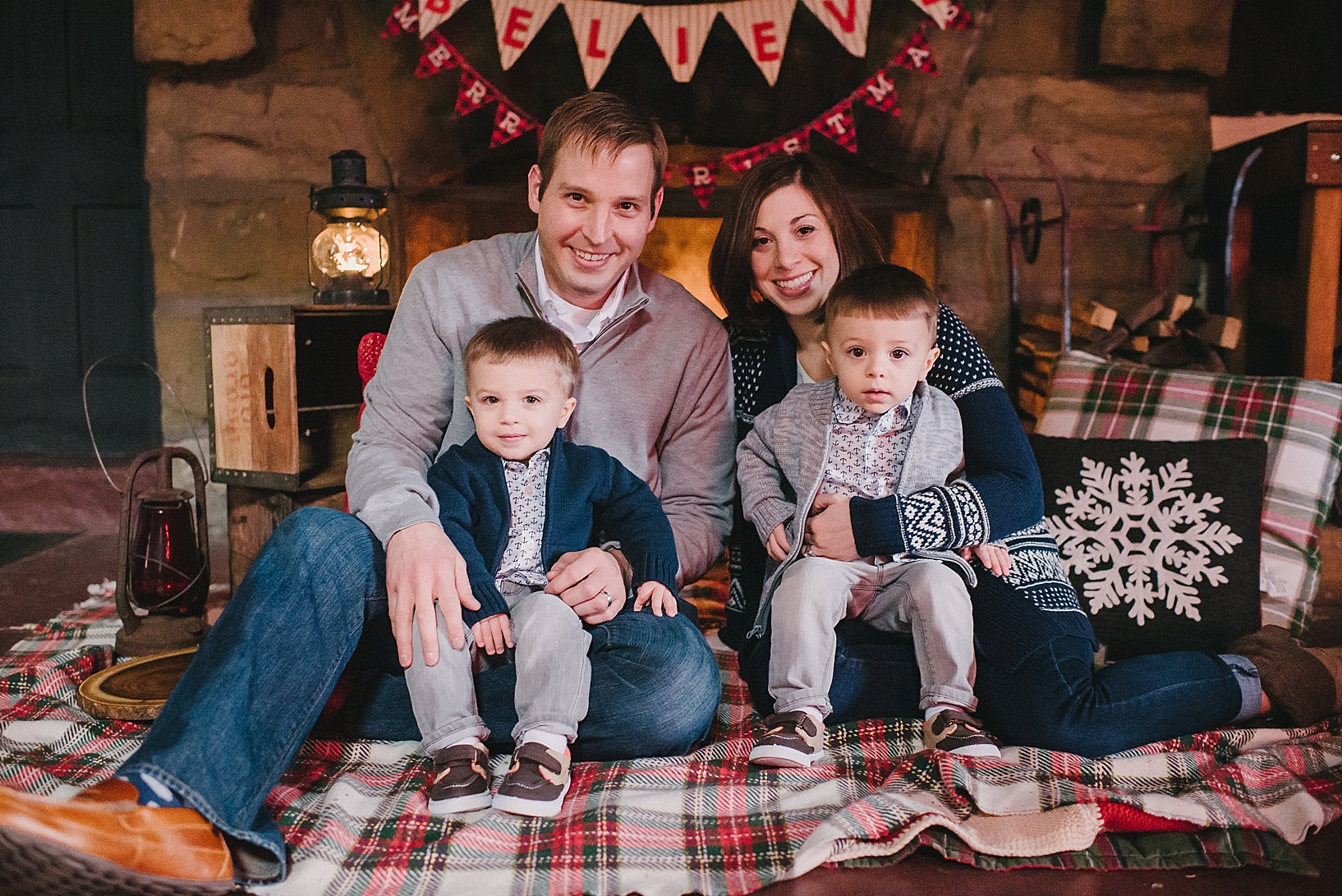 parents with twin toddler boys sitting on plaid Christmas blanket in front of stone fireplace