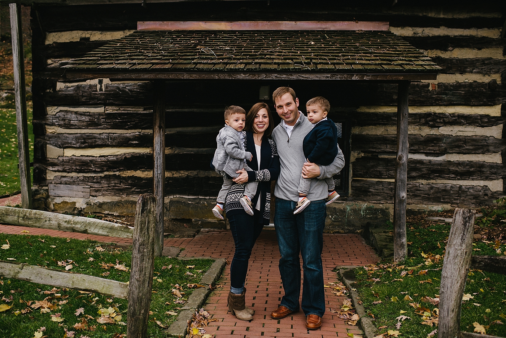 parents holding toddler twin boys standing in front of rustic log cabin wearing navy and grey outfits