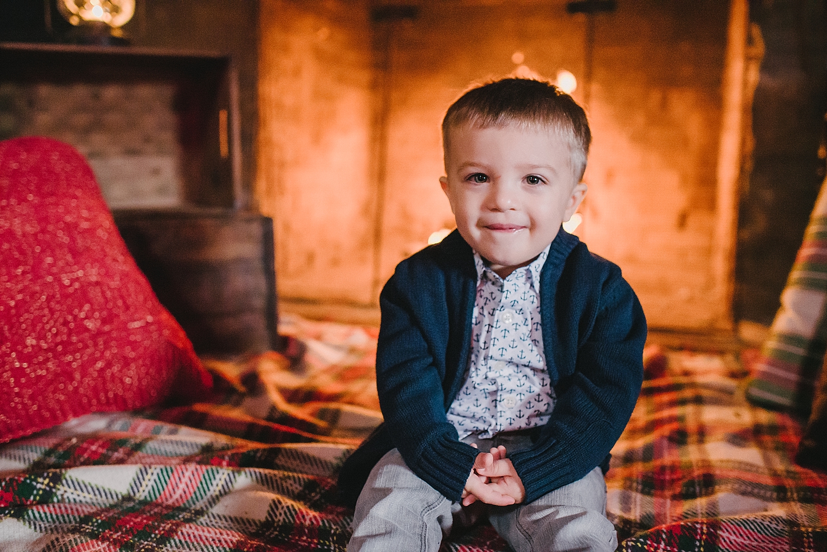 little boy in navy blue cardigan and button down dress shirt sitting in front of crackling fireplace on plaid blanket with hands in his lap