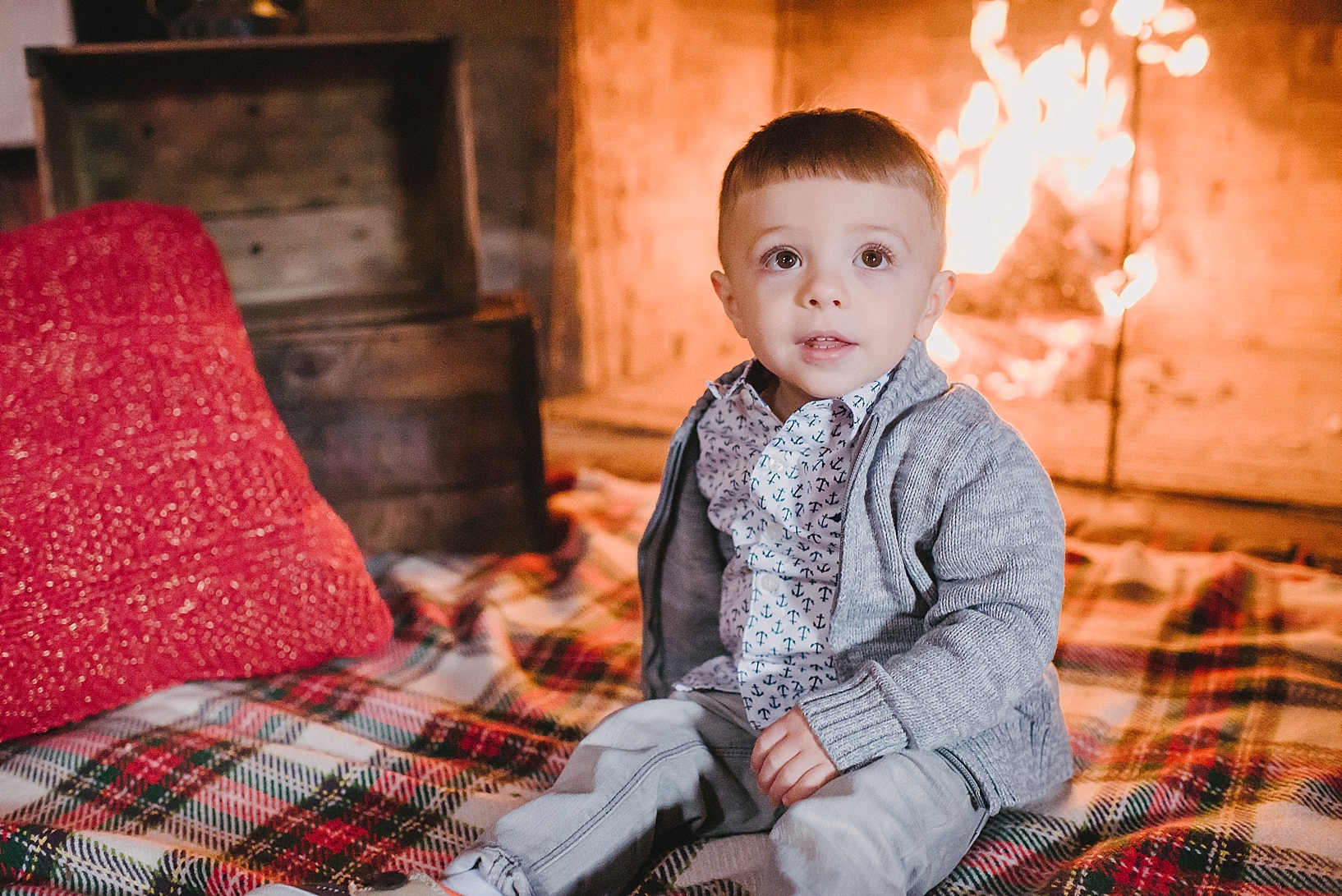 little boy in grey cardigan and button down dress shirt sitting in front of crackling fireplace on plaid blanket