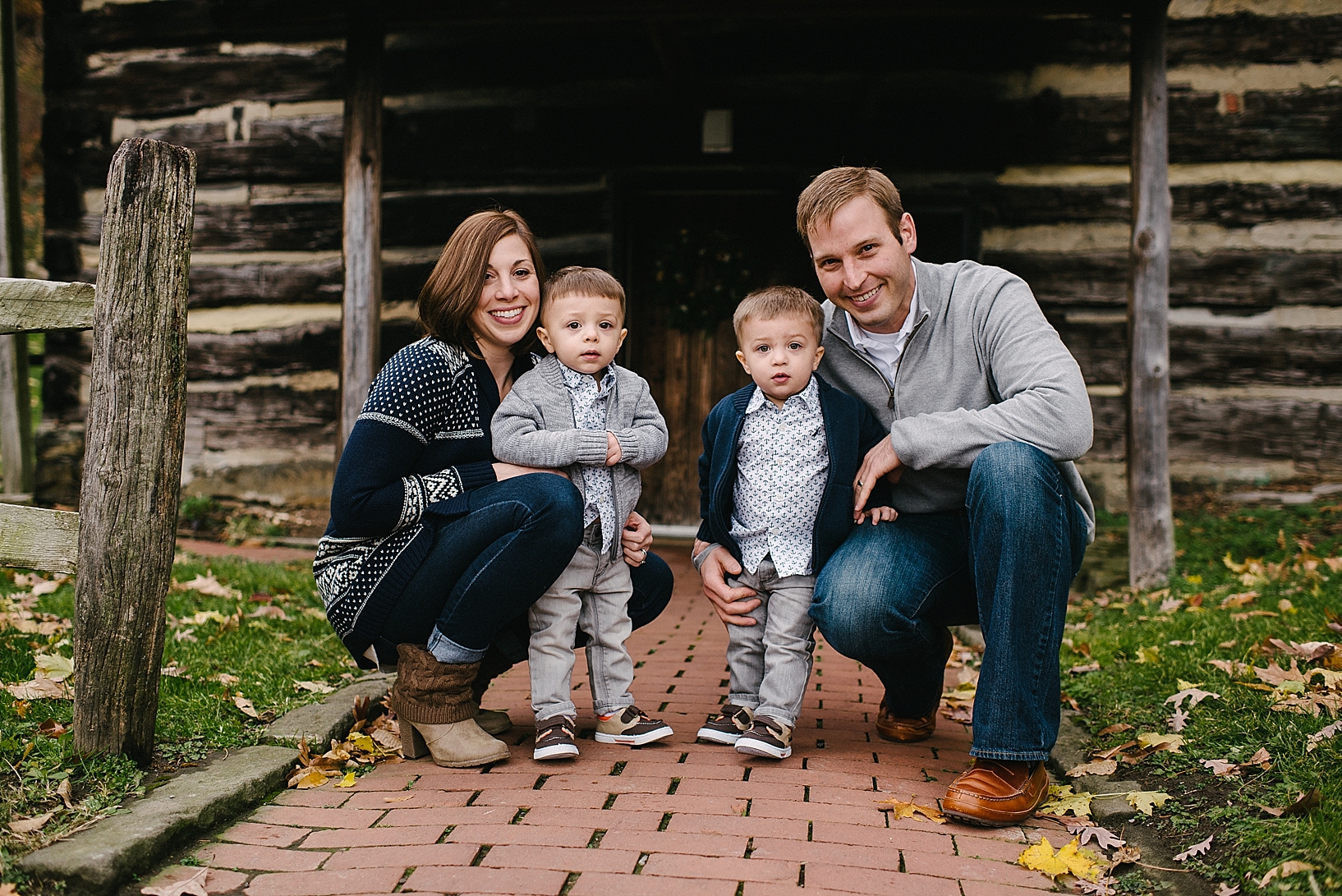 parents with toddler twin boys standing in front of rustic log cabin wearing navy and grey outfits