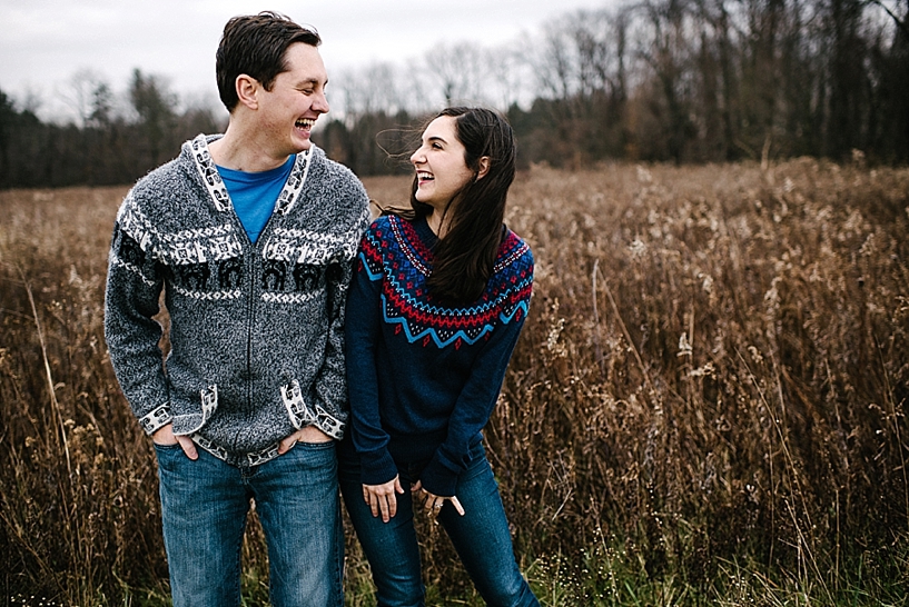 couple wearing winter sweaters standing in field laughing at each other