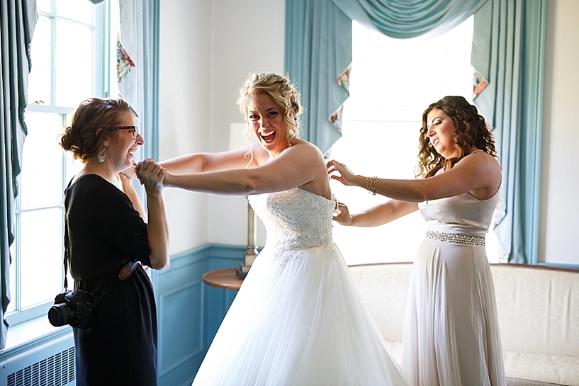 bride holds onto photographer's hands while getting dress laced