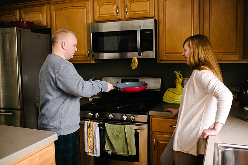 man in grey sweatshirt flips french toast in red skillet as wife looks on smiling