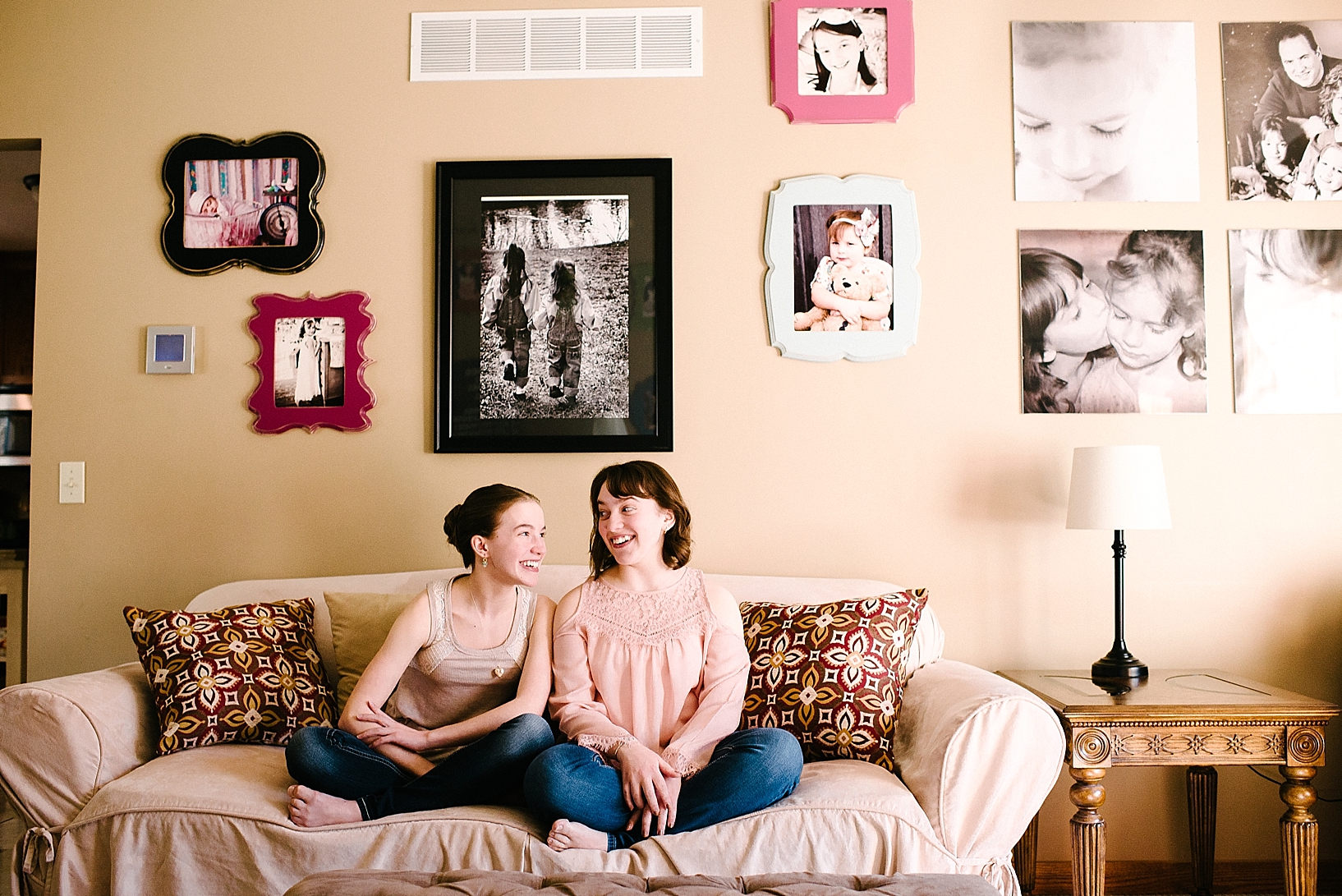 teenage sisters sit cross legged on couch in front of photo wall