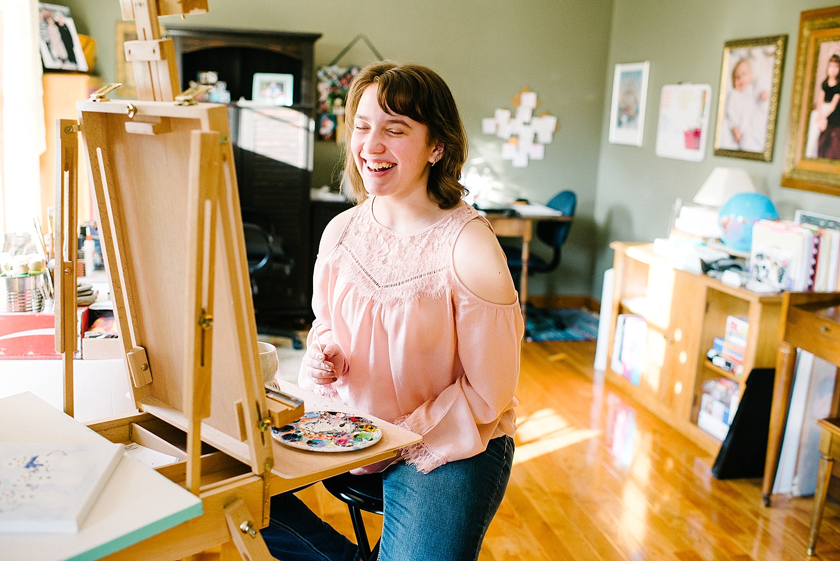 teen girl wearing blush shirt sitting at easel painting and laughing
