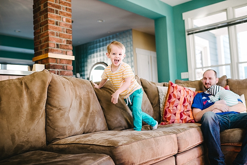 little boy in teal pants jumping on couch with dad holding baby in the background