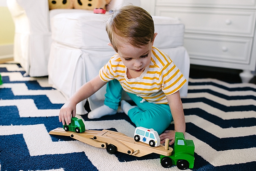 little boy in teal pants and yellow striped shirt playing with wooden trucks on carpet