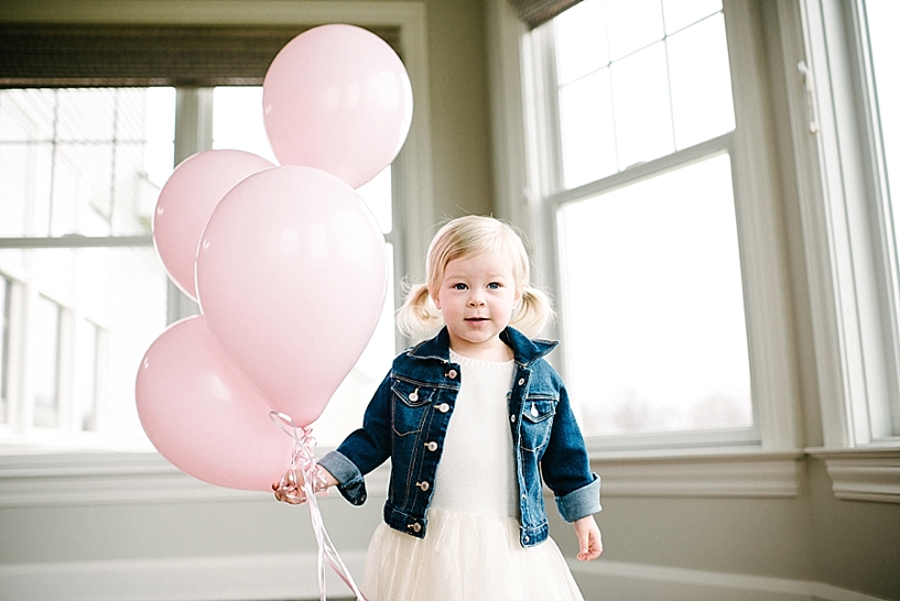 two year old girl wearing dress and denim jacket holding bunch of pink balloons