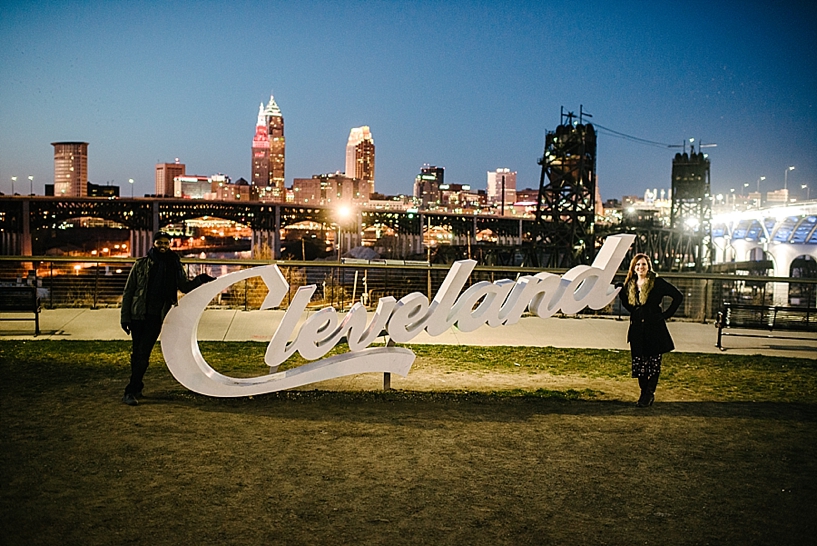 couple standing by white Cleveland sign with skyline in the background at night