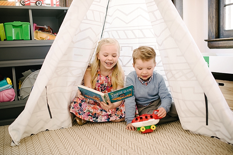 blonde girl in floral dress reading book to blond boy wearing blue sweater sitting in teepee in play room