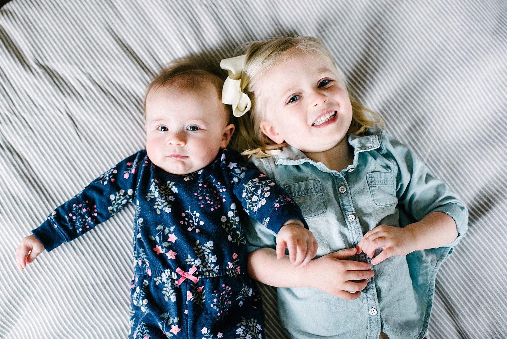 baby girl wearing floral romper and toddler girl with blonde hair wearing denim shirt laying on striped bed sheet
