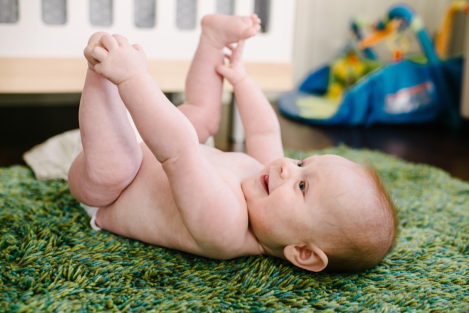 redhead baby boy laying on back on green carpet playing with toes