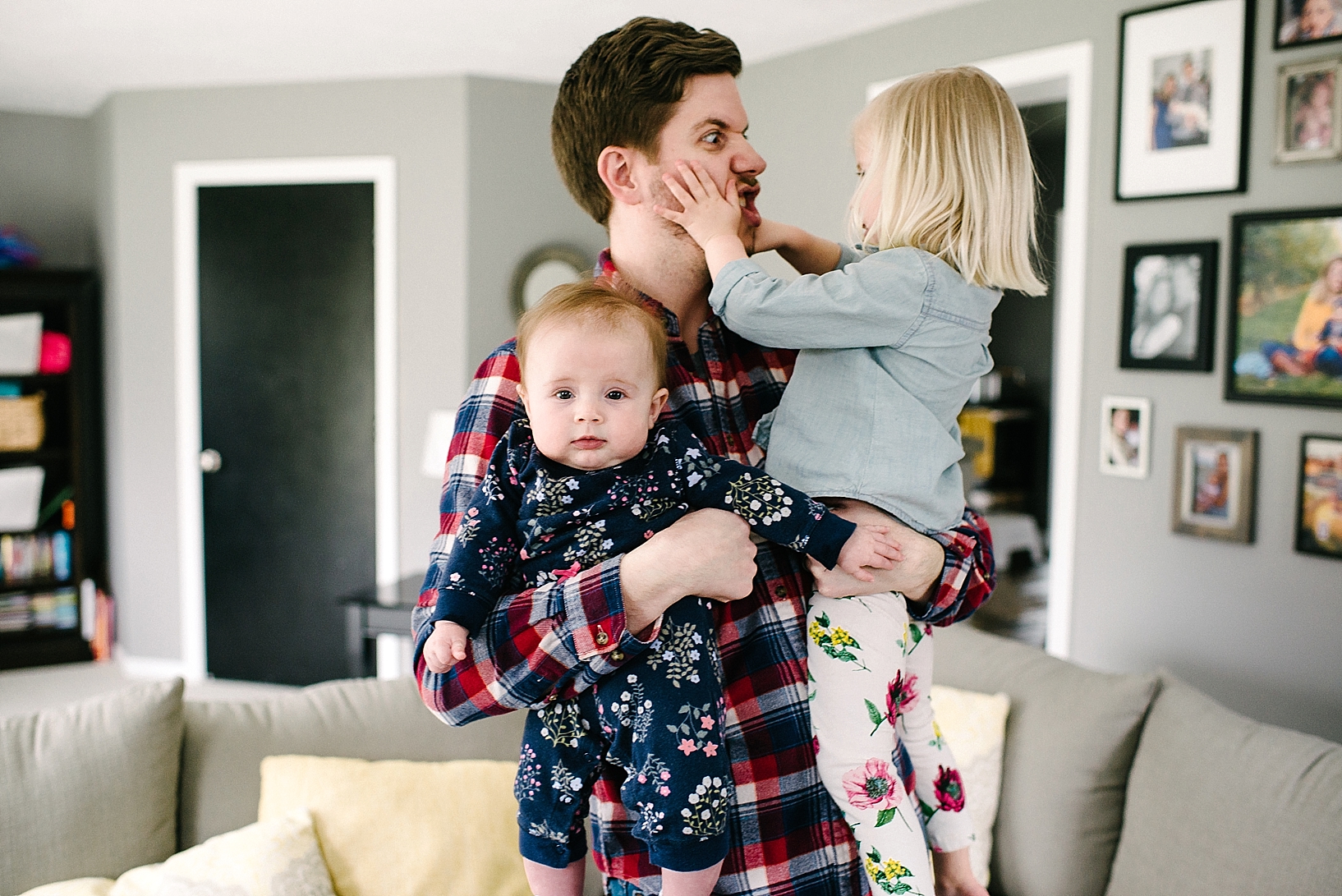 dad standing in living room holding two young daughters in his arms making silly face