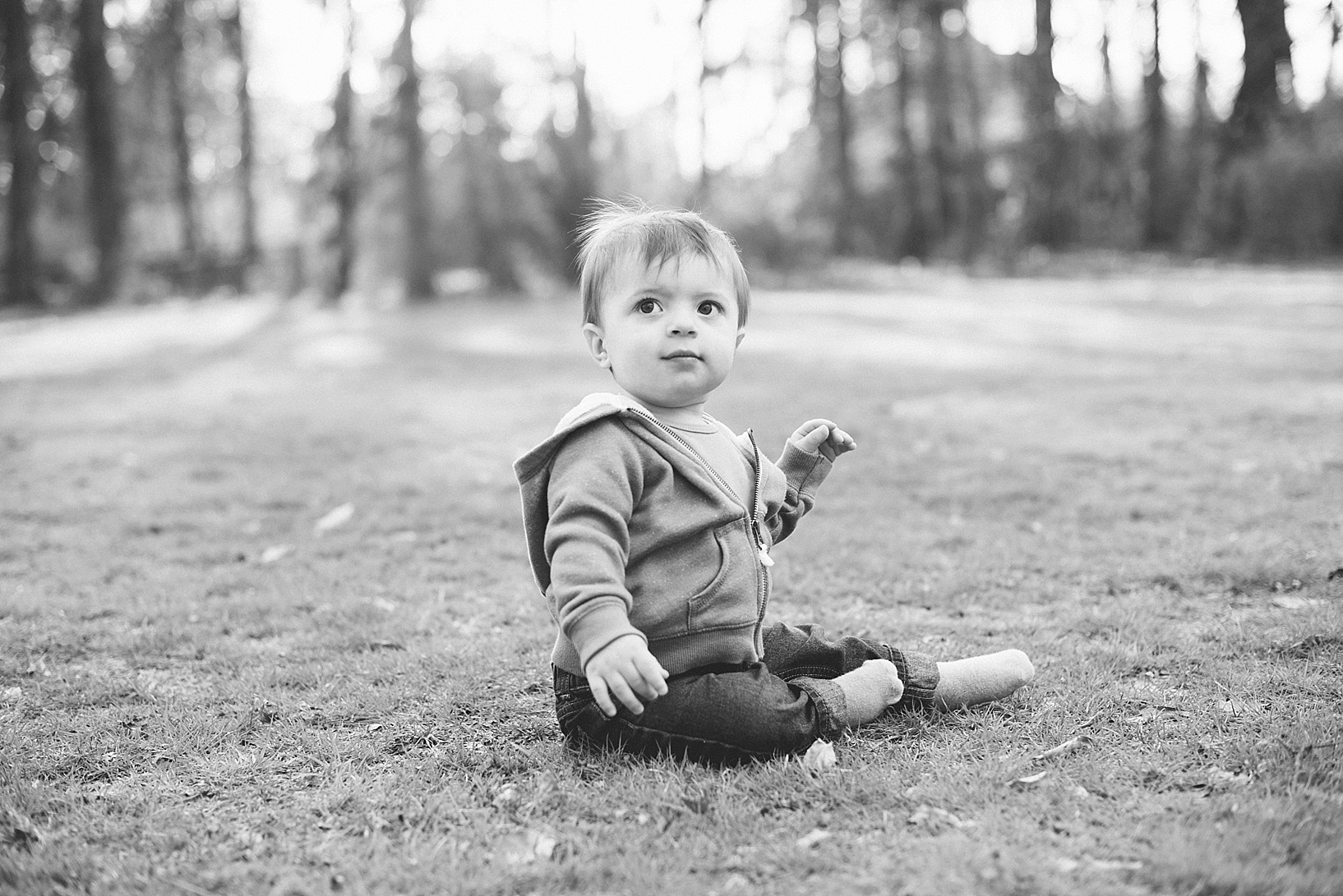 one year old boy wearing hoodie and jeans sitting in grass in the park