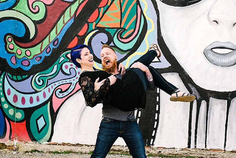 Tattooed man picking up woman with blue hair in front of colorful mural wall