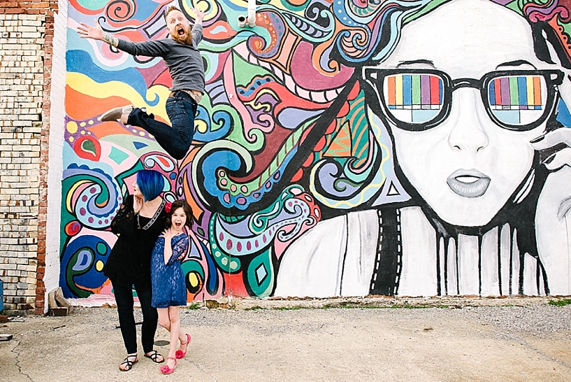 man jumping over wife and daughter in front of mural wall