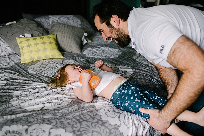 dad laughing at toddler daughter laying on bed