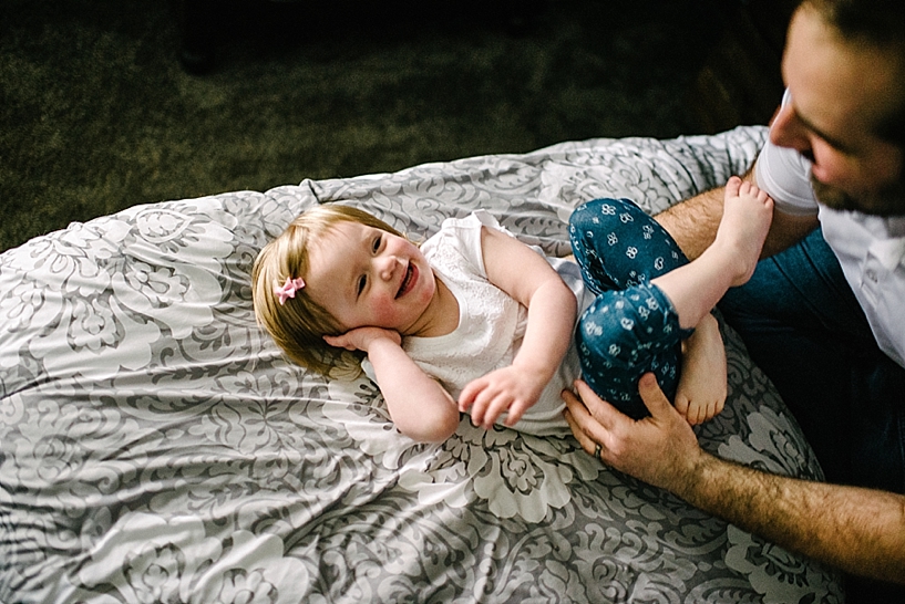 toddler girl laying on grey bedspread smiling up at dad with his arms around her