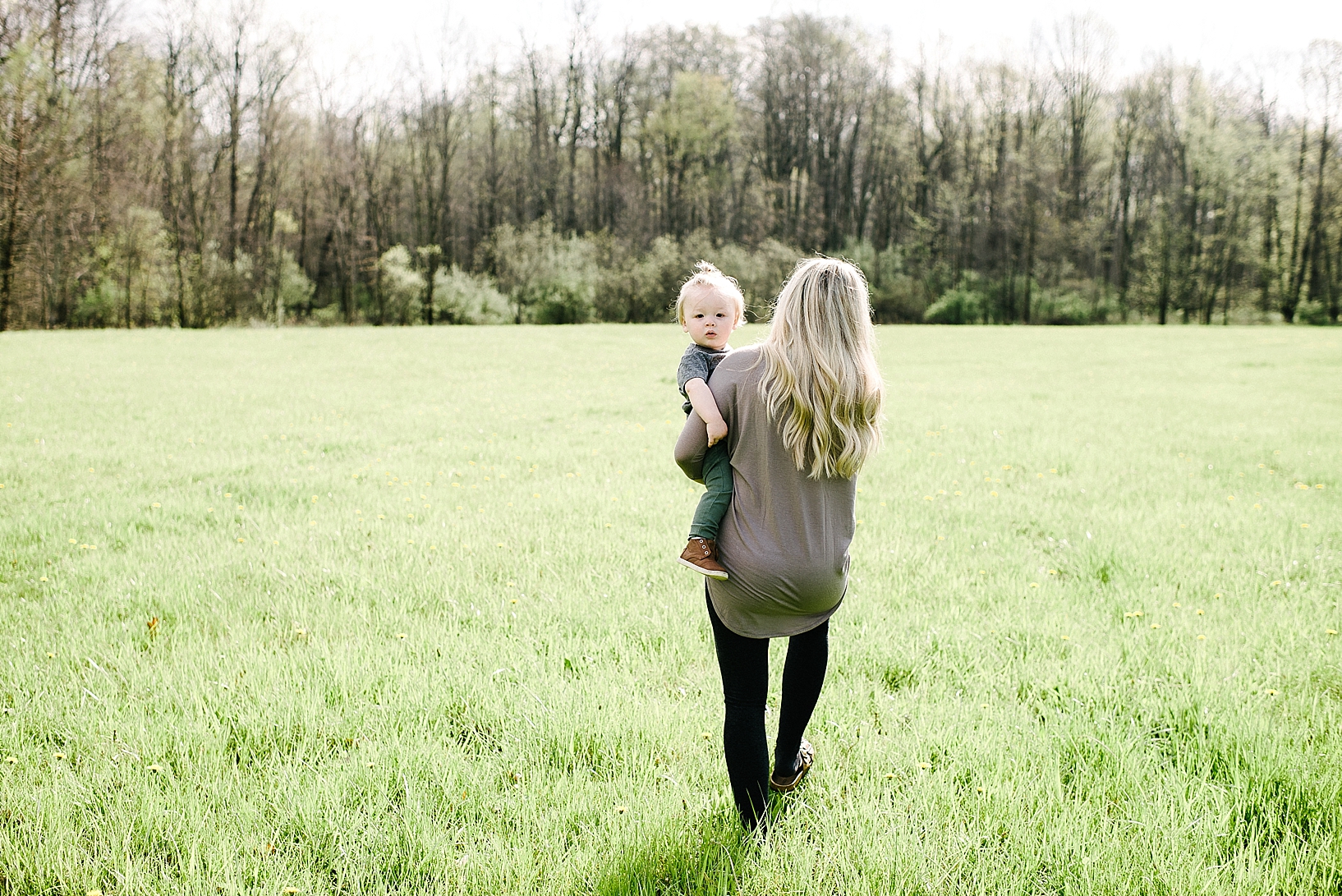 toddler boy with man bun walking across open field being carried by mother with long blonde hair