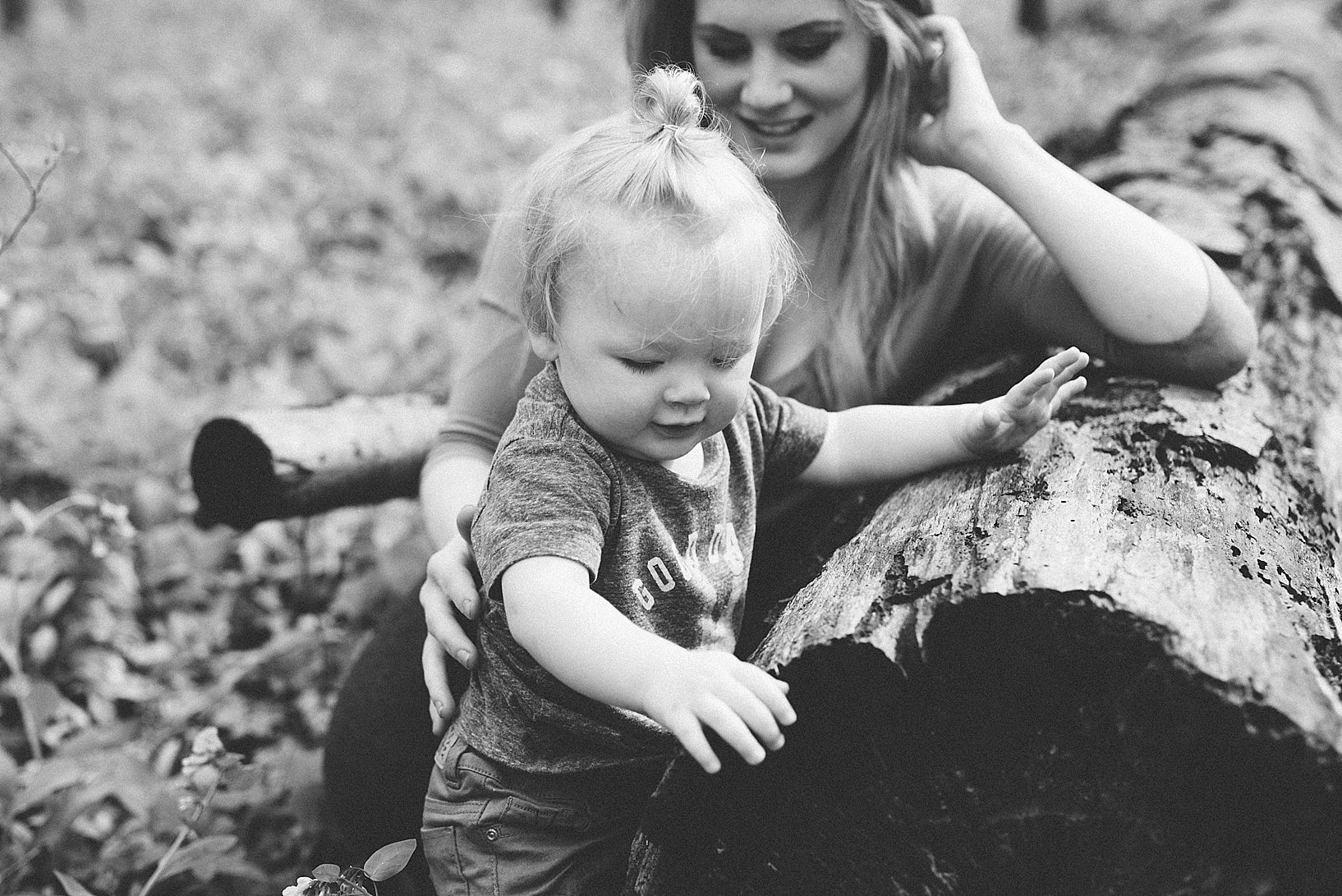 toddler boy with man bun playing on tree log with mother smiling behind him