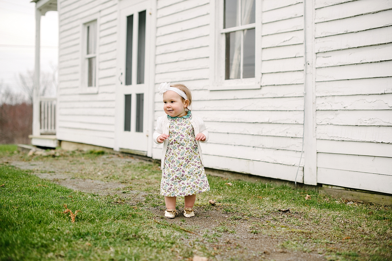 little girl in floral print dress and gold moccasins standing in front of white farmhouse