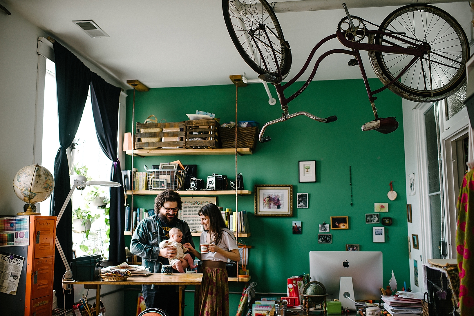 bohemian couple holding baby standing at home studio desk with bike suspended from ceiling