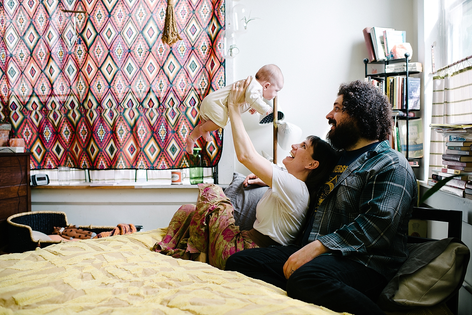 bohemian parents sitting on yellow chenille bed holding baby daughter up in the air