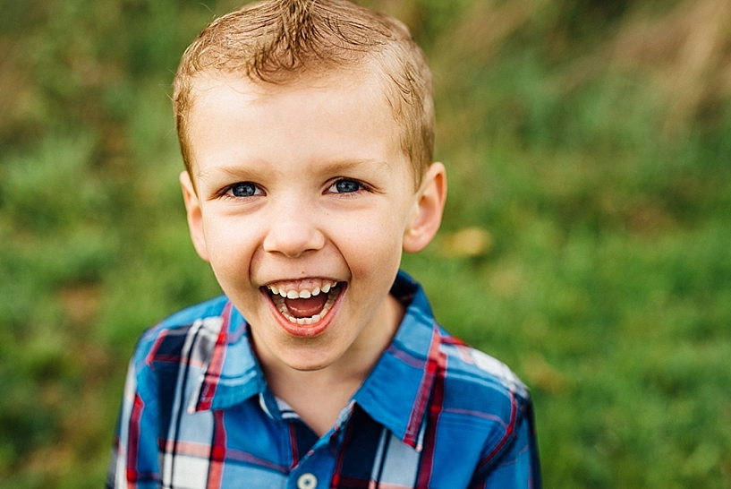 young boy with blue eyes and blonde hair wearing blue and red plaid shirt standing in field of grass