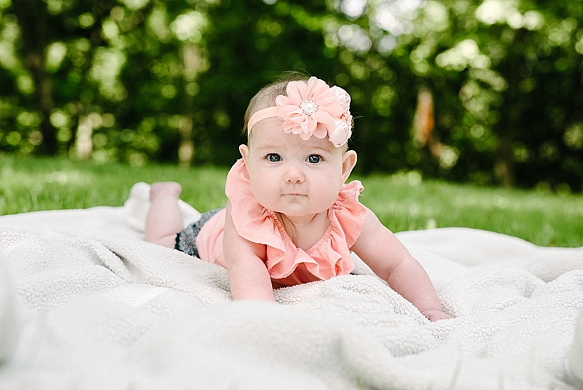 6 month old girl wearing coral shirt with headband laying on blanket in backyard